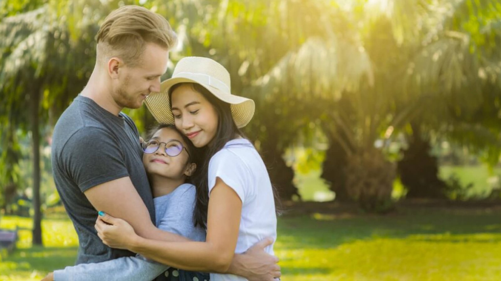 A mother and father embracing their smiling daughter in a sunlit park