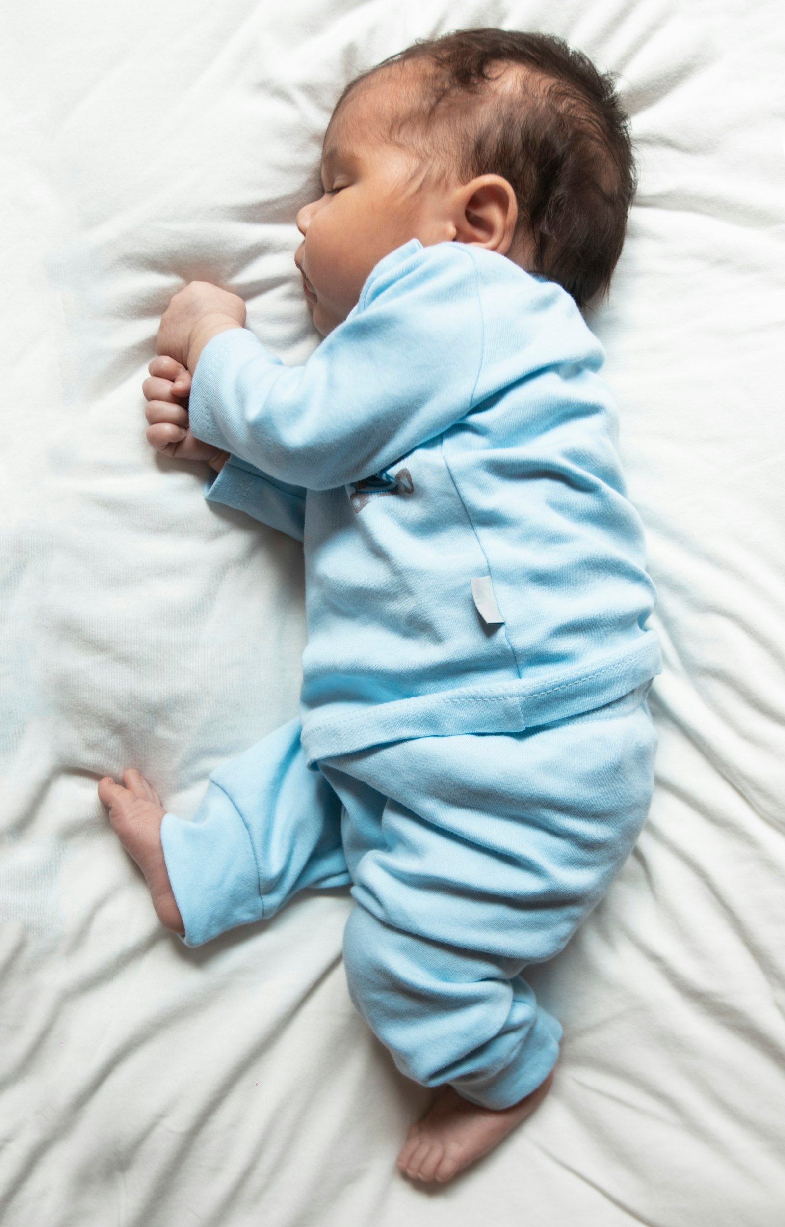 A baby in light blue pajamas sleeping on a white sheet